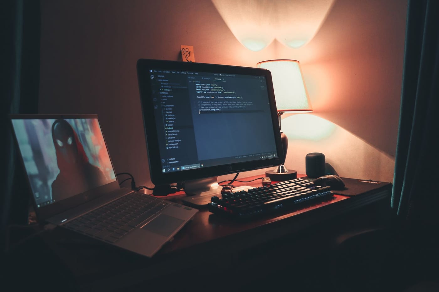 A warmly lit home office at night, two monitors showing code, the room otherwise dark apart from a desk lamp.