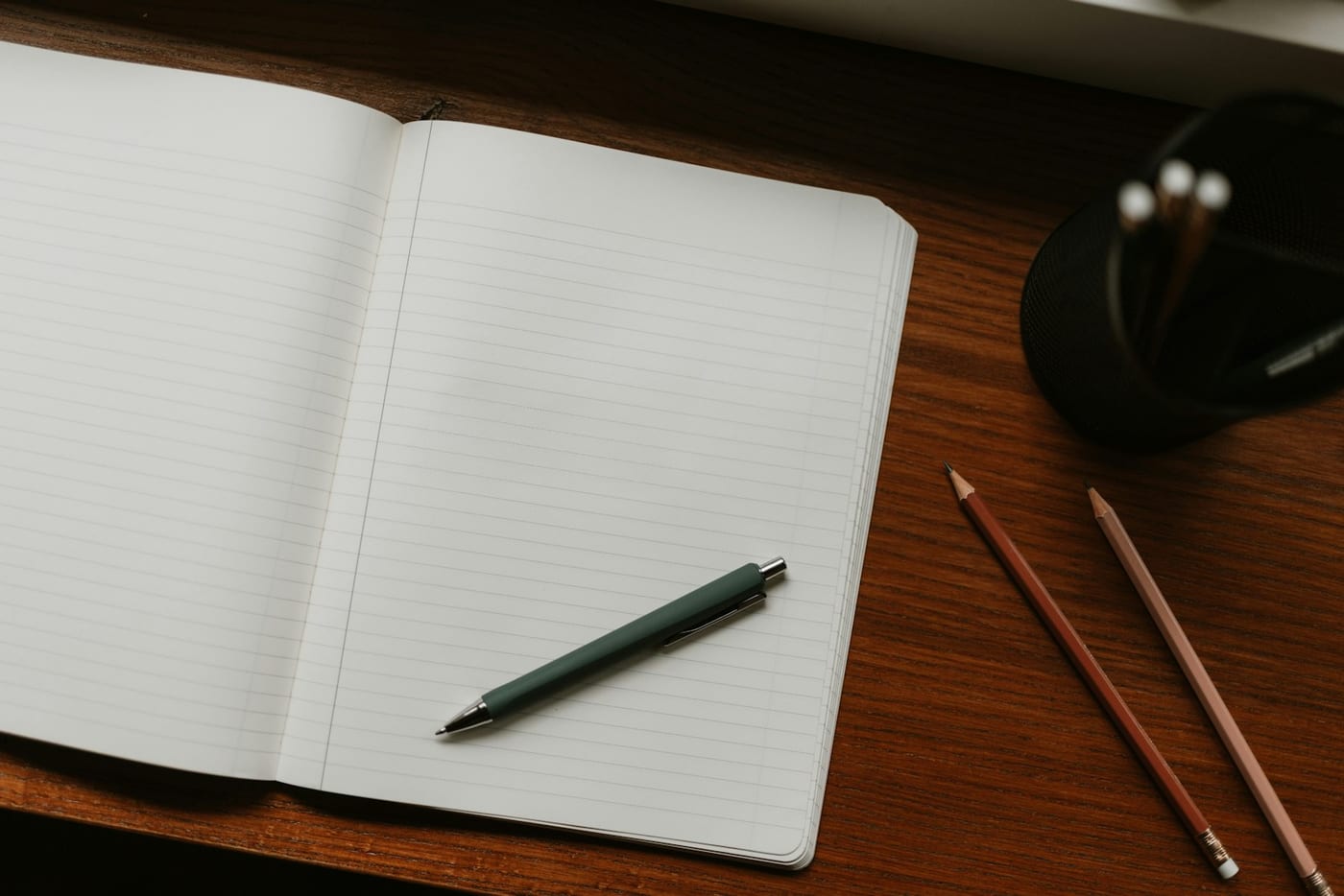 An open notebook with a pen and two pencils laid on a warm wooden desk, the pages blank.