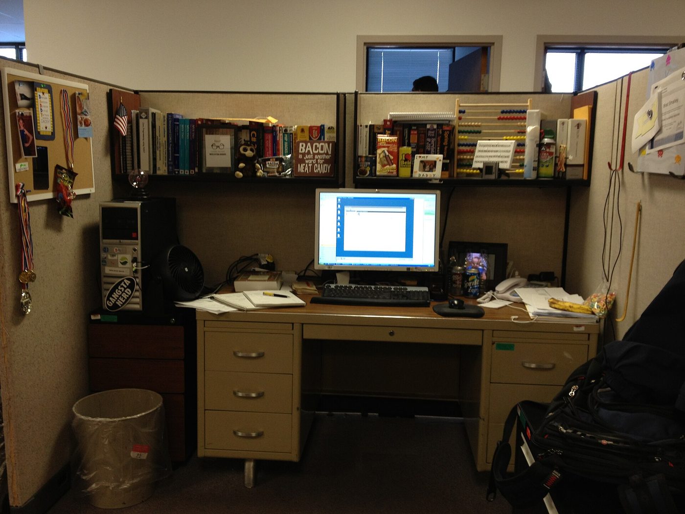 A working programmer's desk in an office cubicle: two monitors, a wall of reference books and manuals, papers, cables and accumulated hardware.