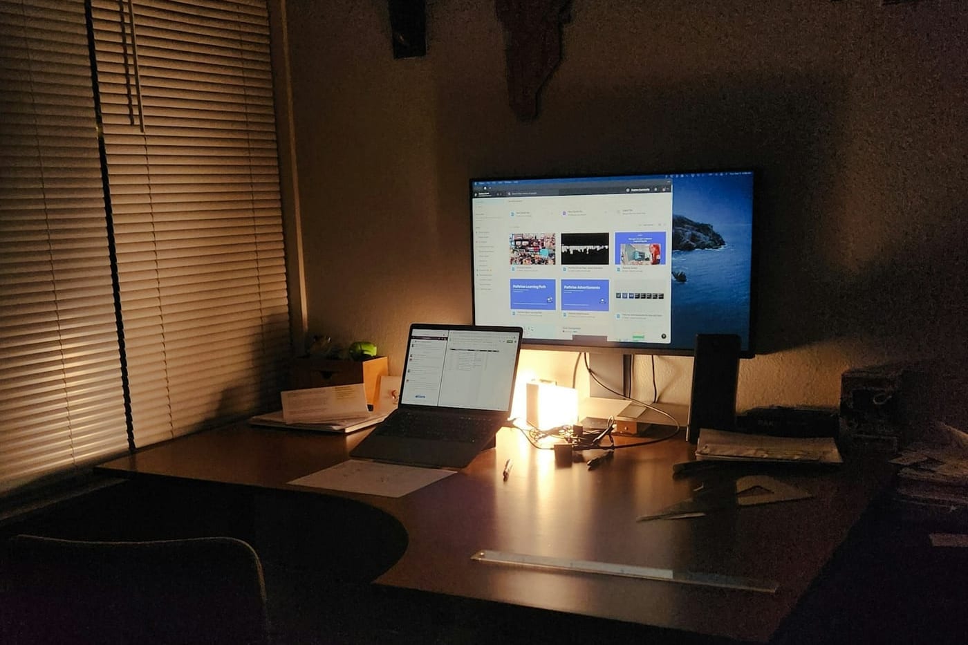 A desk at night lit only by a lamp behind the laptop, a second monitor glowing, the rest of the room in shadow.